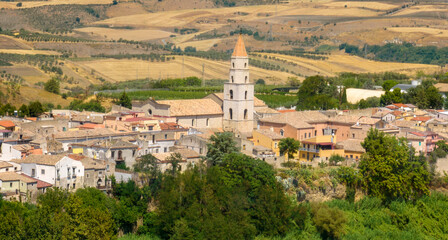 Fototapeta premium Aerial view of a traditional Italian village nestled in rolling hills. A prominent white bell tower with a pointed orange roof rises above the town's historic buildings. It is Venosa, in Basilicata.
