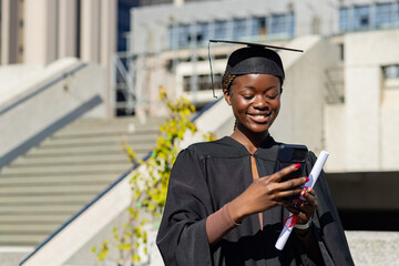 Woman wearing black gown and cap standing on campus stairs, holding diploma, checking phone © wavebreak3