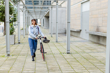 Mid-adult woman walking bicycle through covered corridor on campus, carrying backpack and helmet © wavebreak3