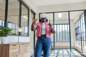 African woman wearing white VR headset, gesturing at virtual content in modern office by dresser © wavebreak3