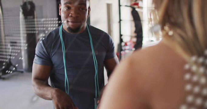Trainer adjusting jump rope instructing client while gesturing down in gym as mirror overlay fading