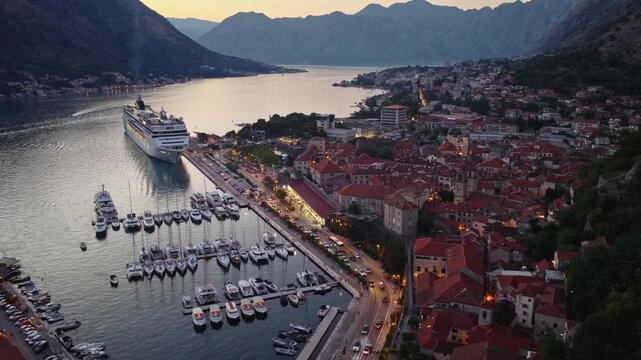Aerial establishing view of Kotor Old town lights in blue hour, mountains range of Kotor Bay fjord in a background, large cruise ship in the port. 