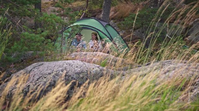 Couple in a green tent among rocks and pine trees in Finland, enjoying peaceful camping in the forest with tall grass in the foreground.