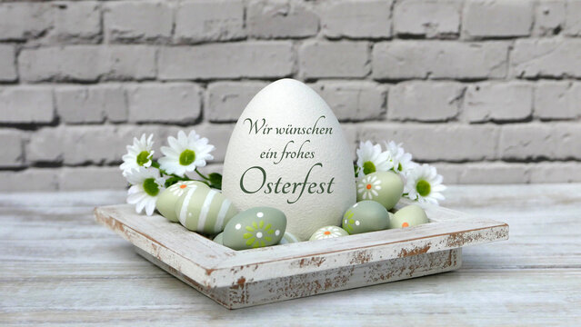 Frohe Ostern:  Osterei mit der Aufschrift "Wir w&uuml;nschen ein frohes Osterfest mit pastellgr&uuml;nen Ostereiern,  und wei&szlig;en G&auml;nsebl&uuml;mchen vor einer Ziegelwand.