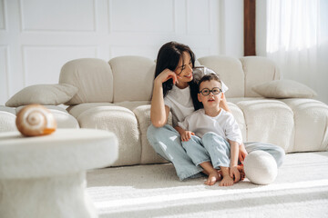 Obraz premium Mother and son sitting on a light-colored sofa in a bright living room, enjoying a moment together with a decorative table and natural light streaming in