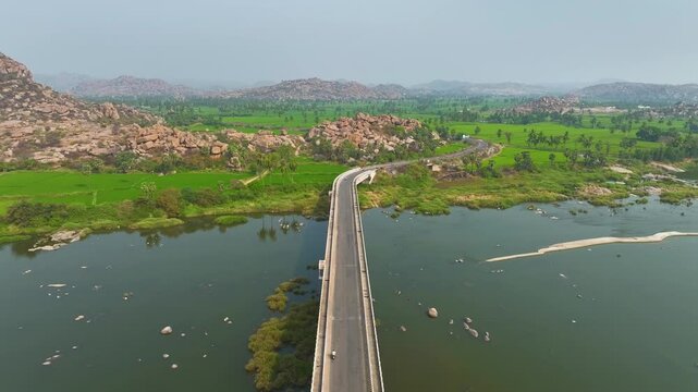 Bridge connecting Hampi villages across Tungabhadra River captured from drone over lush agricultural landscape.
