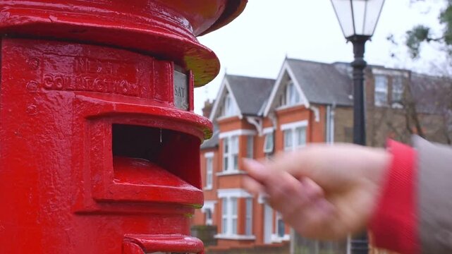 Male hand postcard into red british post box on residential street. Neighborhood classic street scene with traditional mailbox. Concept of communication tradition postal service business