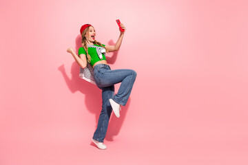 Young woman in bright casual outfit takes a selfie while jumping against pink background expressing...