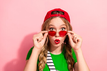 Young stylish woman with red cap green shirt and pink background wearing red sunglass surprised
