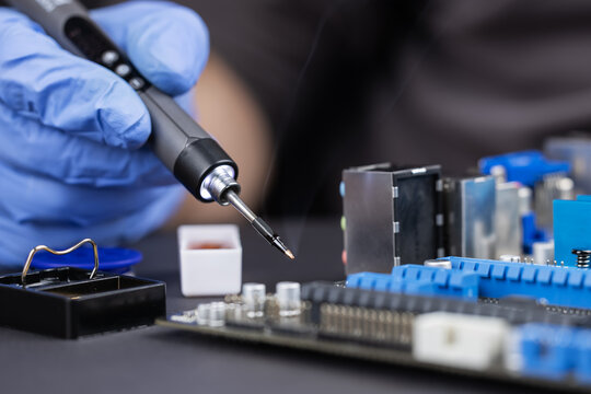 Closeup of worker in protective gloves soldering electronic circuit board with hot iron and visible smoke during assembly process