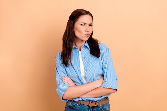 Skeptical young woman with arms crossed wearing a blue striped shirt on beige background portrait expressing doubt and attitude