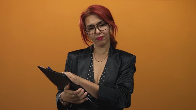 Woman wearing glasses and blazer points pen to clipboard in orange studio while holding notes; concentration professionalism.