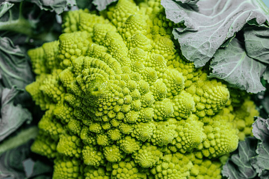 Macro close-up of fresh romaine cauliflower showing detailed spiral fractal florets with natural green texture and surrounding leaves.