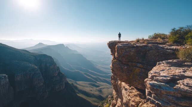 Man standing on edge of a high cliff looking at mountain landscape. Adventure and exploration concept. Traveler in nature during sunny day. Freedom and inspiration in wild environment.