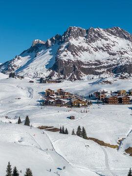 Aerial winter scene of Lech and Oberlech in the Austrian Alps shows chalets, slope side hotels, groomed pistes, ski lifts, resting skiers, evergreens, and a jagged peak.