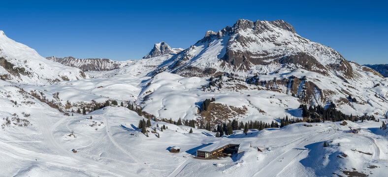 Aerial view of Lech and Oberlech in the Austrian Alps shows groomed pistes, lift lines, chalets, a lodge by dark evergreens, and rugged peaks under bright winter daylight.