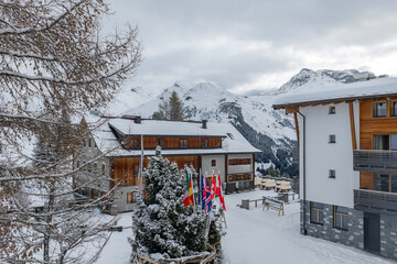 Fototapeta premium Chalet style hotels cluster on a snowy slope in Lech and Oberlech, Austria. Frosted evergreens, Alpine peaks, flags, and terraces with benches mark a calm midday ski season scene.