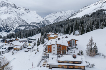 Fototapeta premium Aerial winter view of Lech and Oberlech in the Austrian Alps shows chalets, luxury hotels, groomed pistes, winding roads, and spruce forests under soft overcast light.