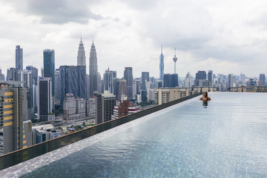 Aerial view of a woman in an infinity pool overlooking the Petronas Towers and Kuala Lumpur Tower against a cloudy sky, Kuala Lumpur, Federal Territory of Kuala Lumpur, Malaysia.
