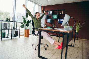 Relaxed businesswoman stretching at her desk in a modern office, smiling and celebrating a moment...