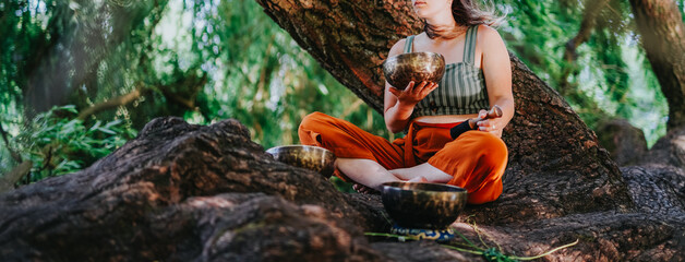 Friends practice a sound bath with Tibetan singing bowls while seated on tree roots. The group...
