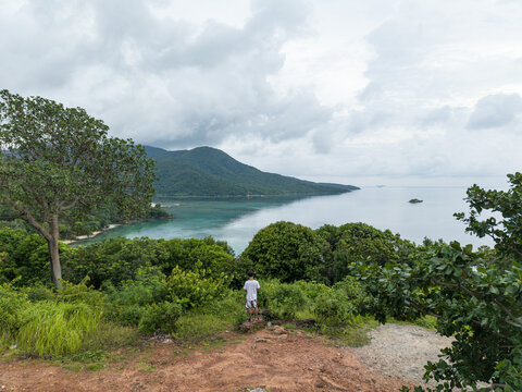 Aerial view of a person standing atop a reddish-brown hill, overlooking the lush green foliage and turquoise sea of Karimunjawa Island, Karimunjawa, Central Java, Indonesia.