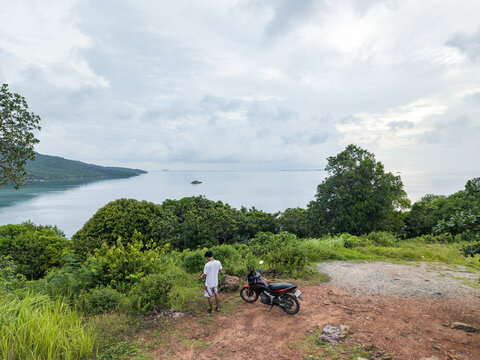 Aerial view of a lone figure stands beside a parked motorcycle, gazing out at the tranquil sea near Cottage Legon Waru, Karimunjawa, Central Java, Indonesia.