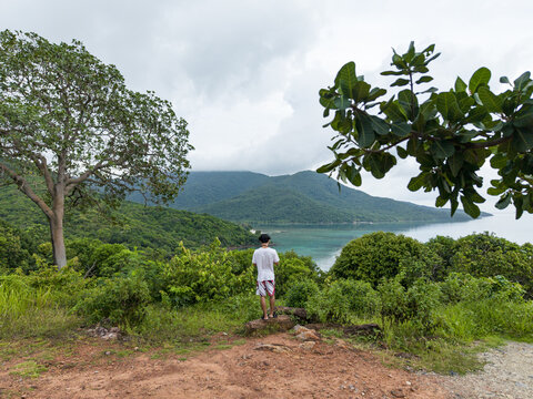 Aerial view of a lone figure standing on a red earth precipice, gazing over the turquoise bay and lush green hills of Cottage Legon Waru, Karimunjawa, Central Java, Indonesia.