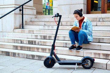 Young adult Caucasian woman sitting on outdoor steps reading book while wearing headphones, electric scooter parked nearby, focusing on studying or relaxing during break