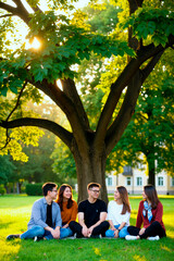 Fototapeta premium Group of five young adult Asian men and women sitting on grass under large tree in park, smiling and talking together, enjoying outdoor leisure time, sunlight filtering through leaves