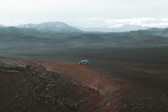 View of a lone car perched on a red ridge overlooking a vast, dark volcanic plain against a backdrop of distant, snow-capped mountains, Rangarthing ytra, Iceland.