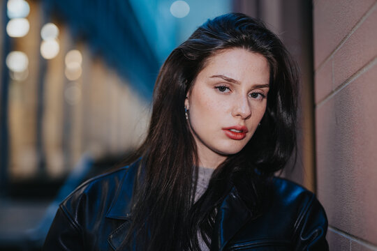Young woman portrait wearing a black leather jacket leaning against a wall. Close-up urban street portrait at dusk with soft bokeh lights in the background.