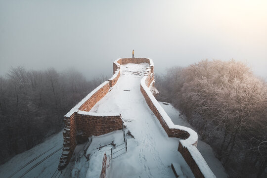 View of the ancient stone fortress, its battlements dusted with fresh snow, rises above the mist-shrouded forest in Chateau du Wegelnburg, Germany.