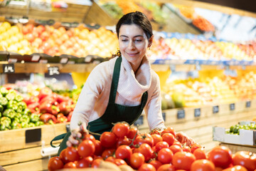 Female seller stands near the counter with red and tomatoes, checks the quality and puts them on the shelf. Supermarket worker keeps an eye on the windows © JackF