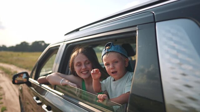 Mother with son looking through car window during family trip. Child waves from vehicle on road journey. Mother and boy enjoy summer travel. Happy family in car explores countryside road trip.