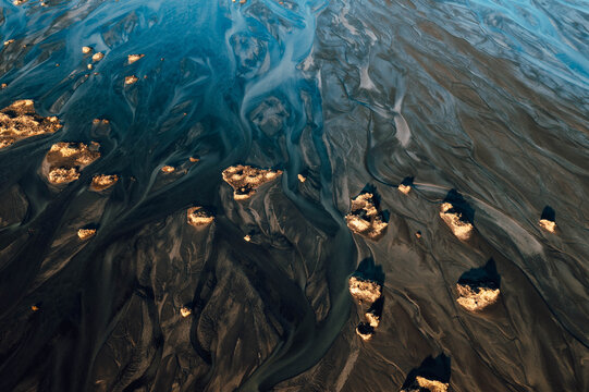 Aerial view of braided river channels carve through the volcanic landscape, creating a tapestry of dark and light hues with golden rocks, Iceland.