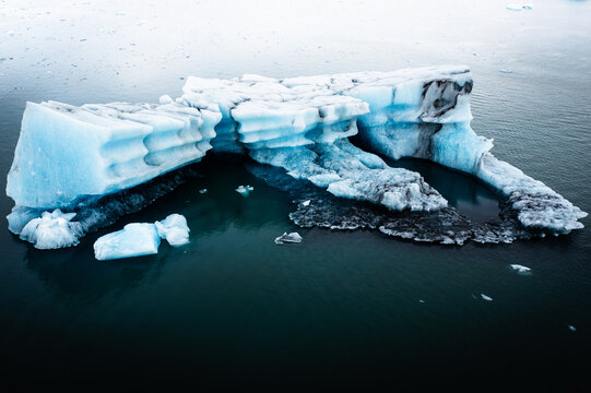 Aerial view of a luminous iceberg drifts serenely in the glacial lagoon, its icy blue hues contrasting with the dark, reflective waters, Jokulsarlon, Iceland.
