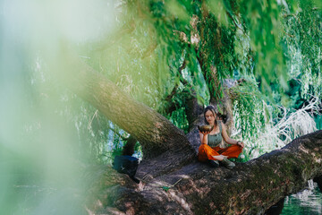 Fototapeta premium Girl meditating on a wide tree branch holding a singing bowl with eyes closed. A peaceful outdoor meditation scene beneath hanging willow branches by the water.