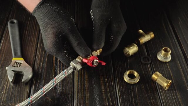 Technician in black gloves tightens a valve connection with a wrench, surrounded by brass fittings and a flexible hose on a wooden work surface
