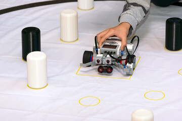 A child's hand interacts with a modular robot equipped with sensors on a white practice mat with obstacles. The image highlights hands-on learning, robotics education for kids, and the development of  © ZeDesign