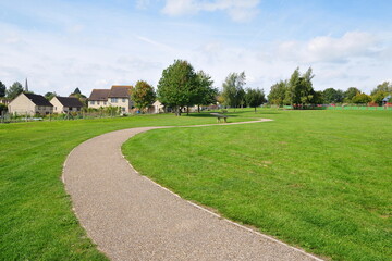 Naklejka premium A winding gravel footpath meanders through a vibrant green park with lush grass recreation field, leafy trees and blue sky above