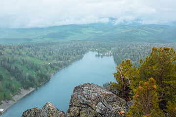 Obraz premium Top view from rocky precipice edge with coniferous trees to big wood lake against hilly vastness far away in low clouds. Conifer grove on rock cliff with view to long alpine lake under cloudy sky.