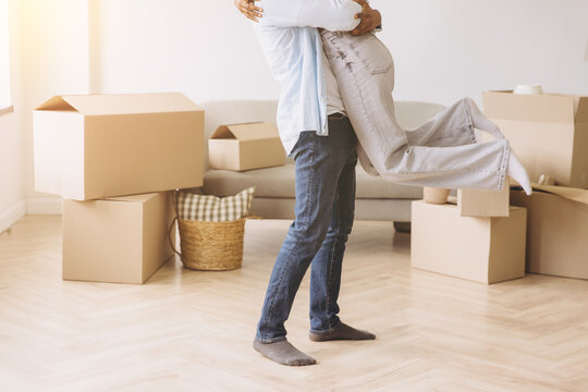 Young couple celebrating moving day in new apartment. Man lifting woman, hugging happily among cardboard boxes