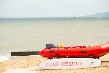 A red inflatable motorboat and a white surfboard with surf rescue text sit on a sandy beach by the sea under a clear sky.