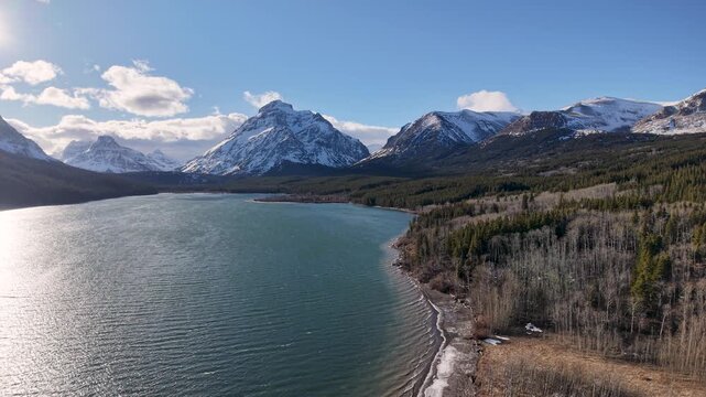 Drone footage captures a wide mountain lake with emerald water, bordered by a forested shoreline and towering snow‑capped peaks under a blue sky near Whitefish, Montana.