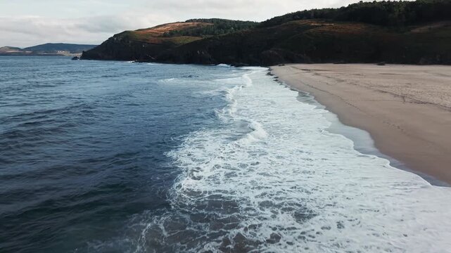 Low drone flying forward above waves along Praia do Rostro beach in Galicia, Spain approaching shoreline with coastal hills and vegetation.