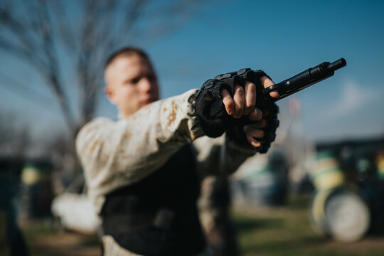 Man wearing gloves and a vest points a paintball gun forward in an outdoor field. The scene emphasizes action and focus with a blurred background.