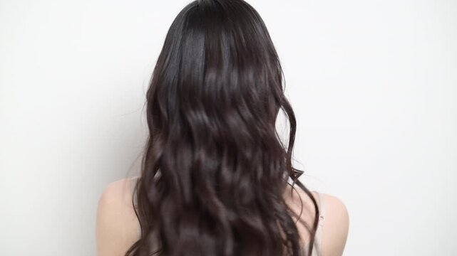 A woman with long, wavy hair and beautiful black locks against a white background; a video showing hair care and treatment techniques, featuring a faceless figure seen from behind; in slow motion