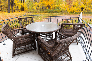 Fototapeta premium Wicker patio table and chairs arranged on a balcony with vibrant yellow autumn trees beyond, suggesting cozy outdoor dining, relaxation and a peaceful seasonal atmosphere