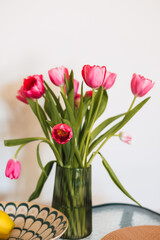 Fototapeta premium Pink tulips in a green ribbed glass vase on a table. Vibrant spring flowers, minimalist home decor, bright natural light, floral bouquet, and fresh aesthetic lifestyle photography. Bulb flower blooms.